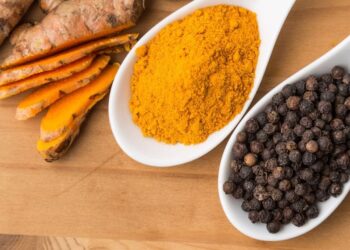 Turmeric powder and black peppercorns on white spoons next to fresh turmeric roots on a wooden surface.