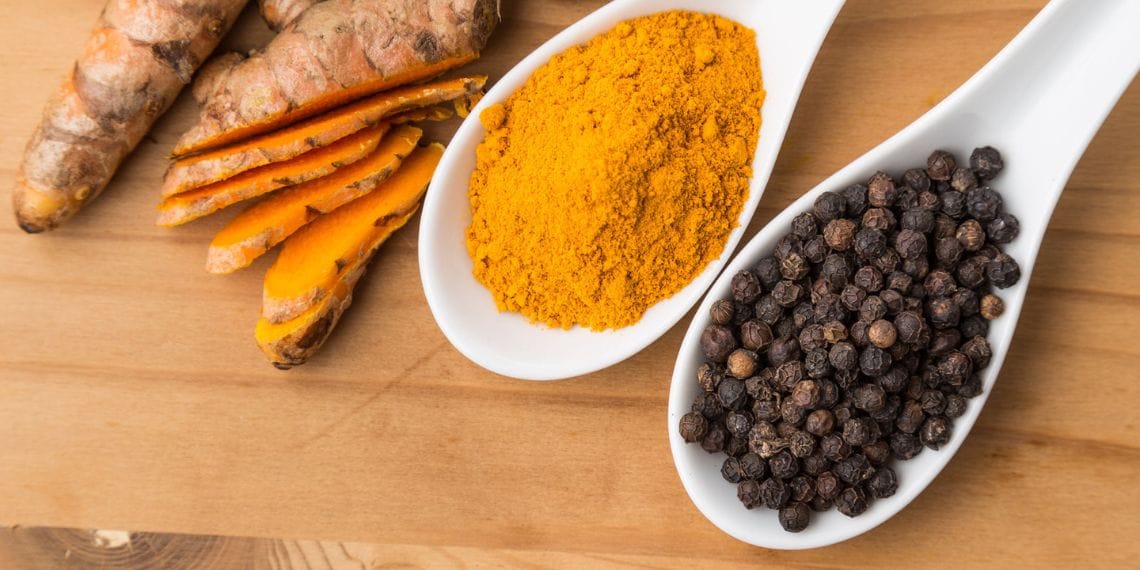 Turmeric powder and black peppercorns on white spoons next to fresh turmeric roots on a wooden surface.