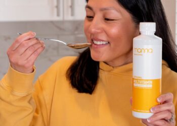 A woman smiling while preparing to take liquid from a spoon, holding a bottle labeled as a multivitamin supplement.