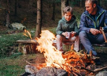 A man and a child sitting on a log by a campfire in the woods.