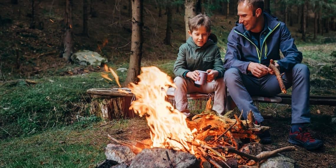 A man and a child sitting on a log by a campfire in the woods.