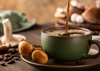 Coffee being poured into a green cup with coffee beans and mushrooms on the table.