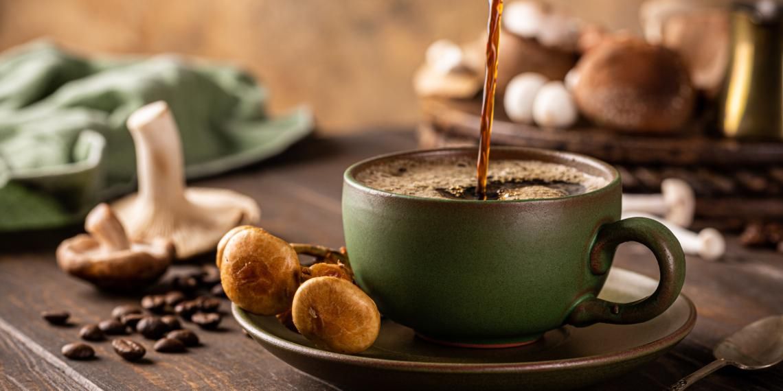 Coffee being poured into a green cup with coffee beans and mushrooms on the table.