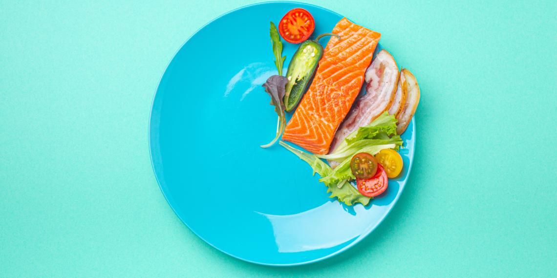 A plate with a raw salmon fillet and fresh vegetables on a turquoise background.