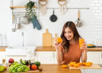 A woman smiling in a kitchen, holding a glass of orange juice and an orange, with fresh fruits and vegetables on the counter.