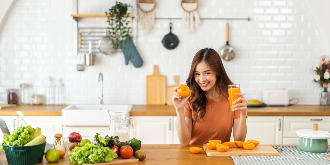 A woman smiling in a kitchen, holding a glass of orange juice and an orange, with fresh fruits and vegetables on the counter.