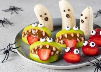 Healthy fruit snacks creatively designed to look like monsters and ghosts for halloween, decorated with candy eyes and displayed on a plate with decorative spiders around.