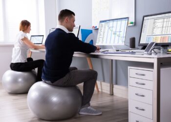 Two office workers using computers while sitting on exercise balls at desks with multiple monitors displaying data.