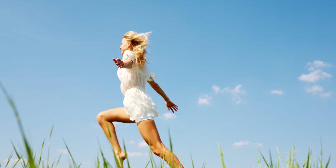 A person in a white outfit is leaping joyfully in a field of grass under a clear blue sky.