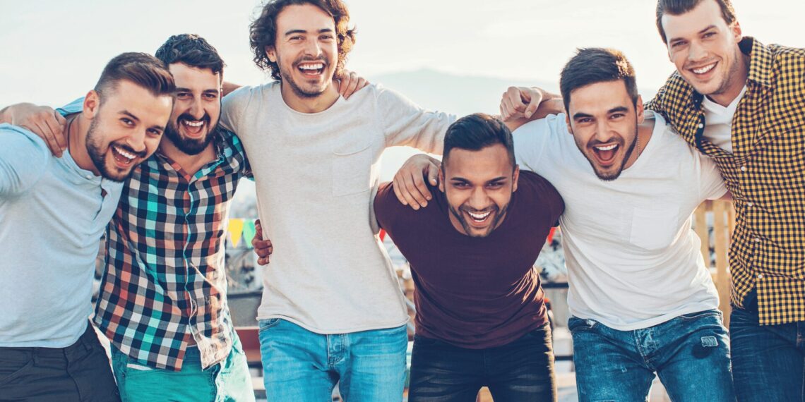 Six men are standing in a row with their arms around each other, smiling and posing for a group photo outdoors.
