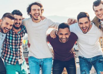 Six men are standing in a row with their arms around each other, smiling and posing for a group photo outdoors.