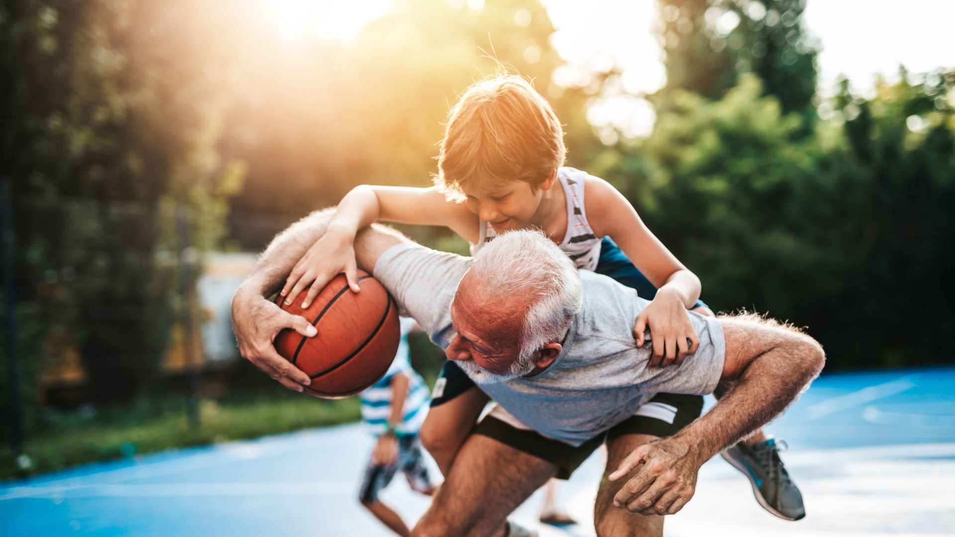 An older man carrying a boy on his back holds a basketball while bending over on an outdoor court with a blurred background of trees and sunlight.