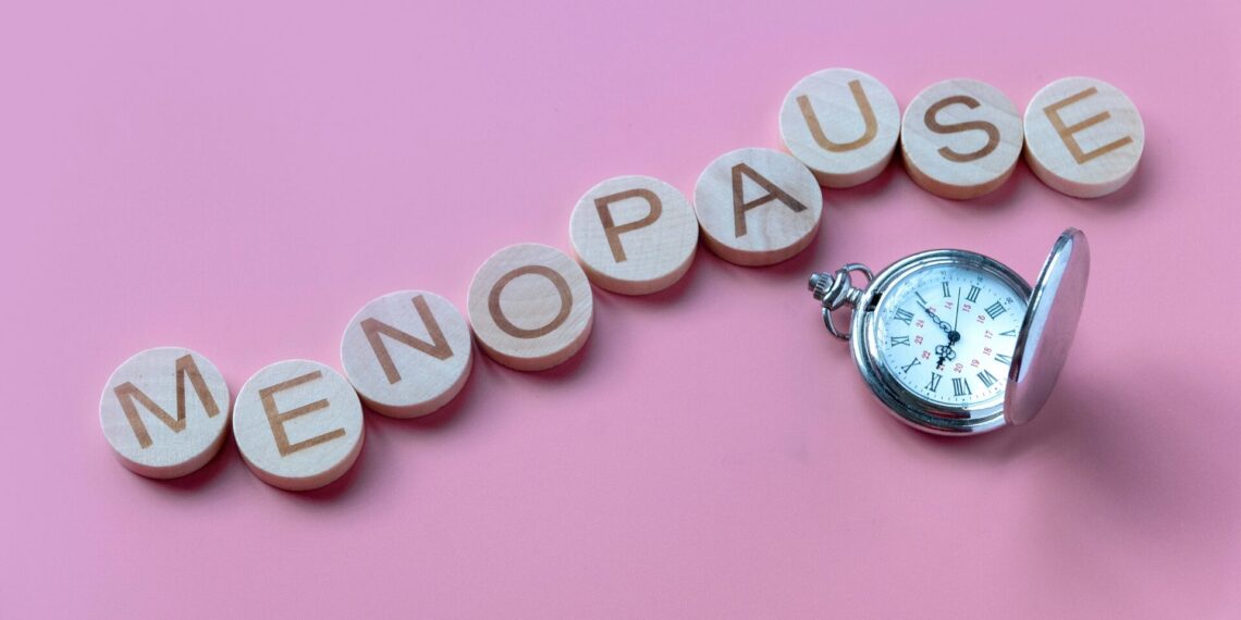 Wooden blocks spelling "MENOPAUSE" arranged in a line with an antique pocket watch next to them on a pink background.