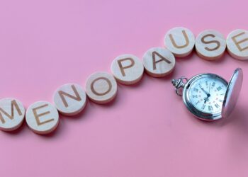 Wooden blocks spelling "MENOPAUSE" arranged in a line with an antique pocket watch next to them on a pink background.