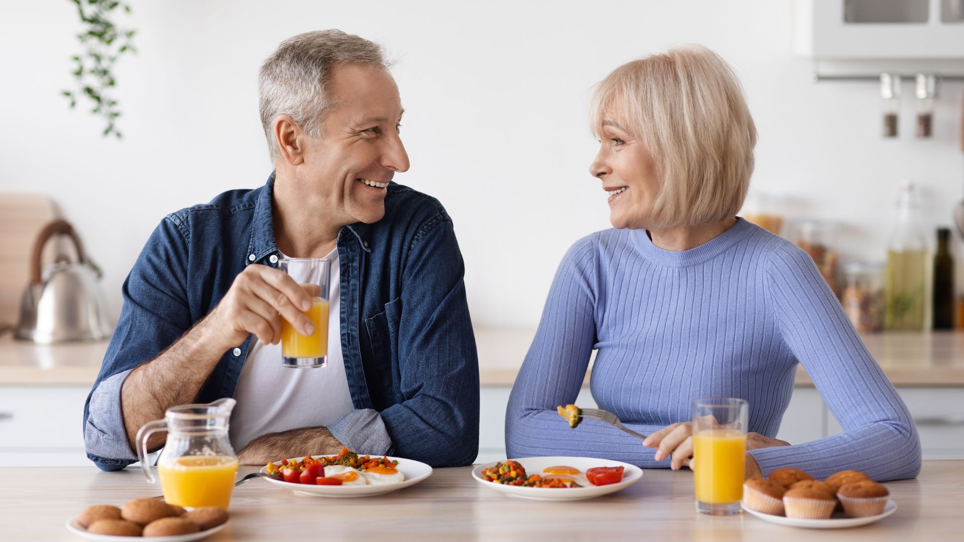 An older couple sitting at a kitchen table, smiling at each other while having breakfast with orange juice and assorted food.