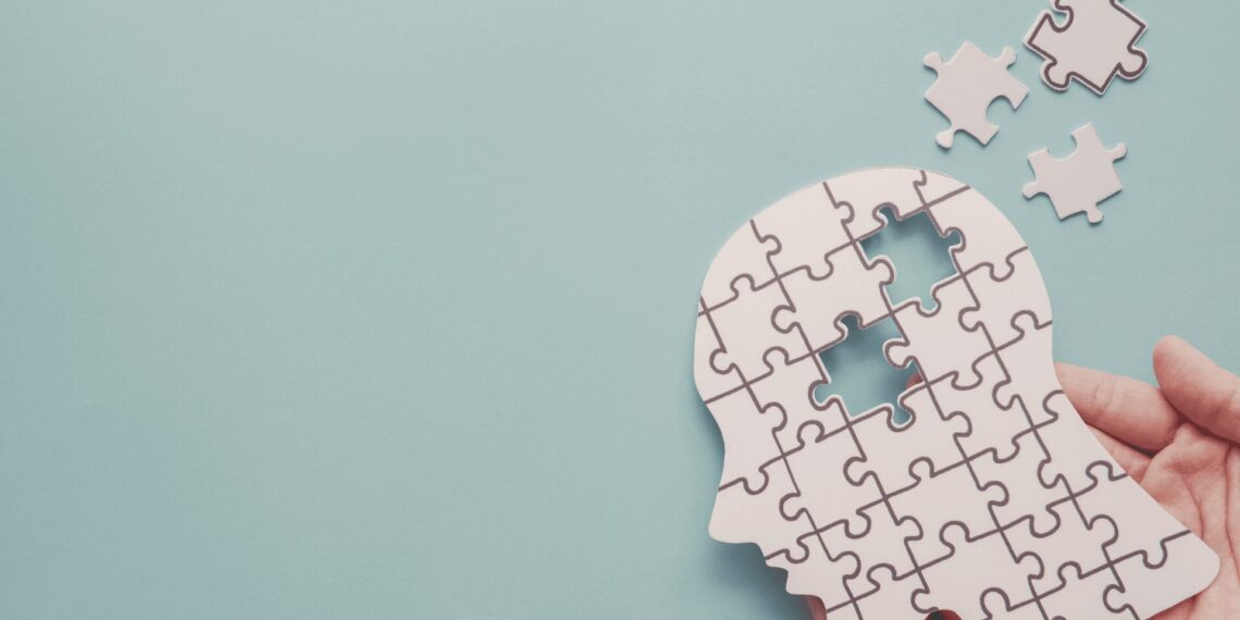 A hand is holding a white puzzle in the shape of a human head with a few missing pieces against a light blue background.