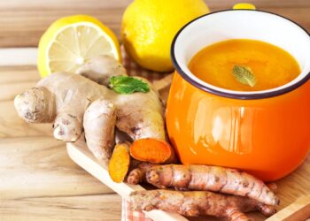 A wooden tray holds fresh ginger and turmeric roots, half a lemon, a whole lemon, and a vibrant orange mug filled with a yellow liquid, garnished with a mint leaf.