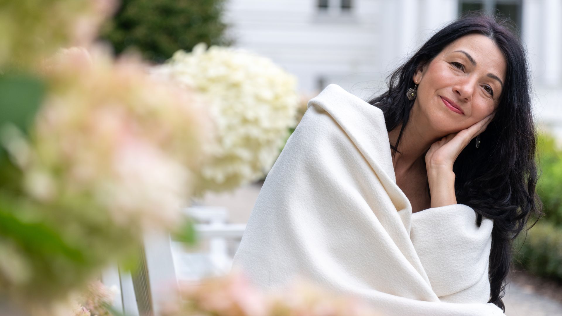 A woman with long dark hair sits outdoors, wrapped in a white blanket, resting her cheek on her hand, with flowers and greenery blurred in the background.