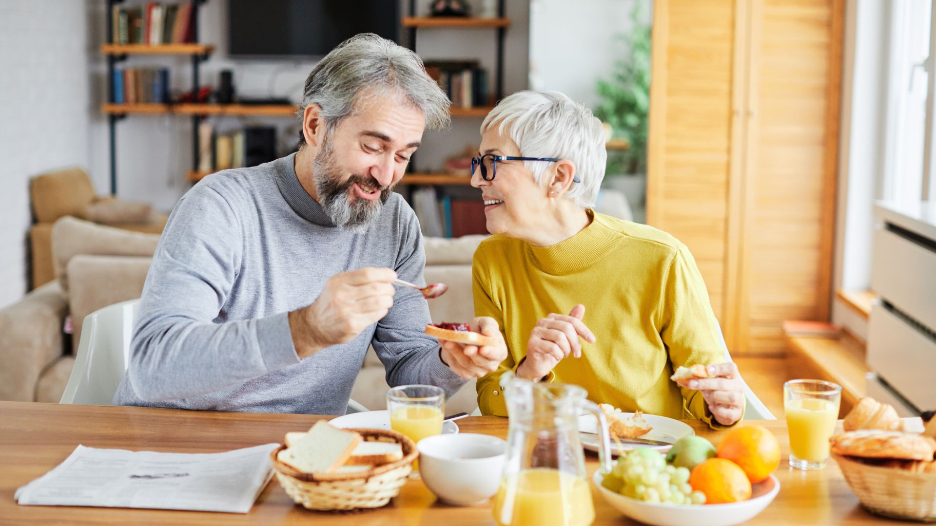An older man and woman sit at a table enjoying breakfast together. The table has bread, fruit, orange juice, and a newspaper. Both are smiling and chatting.