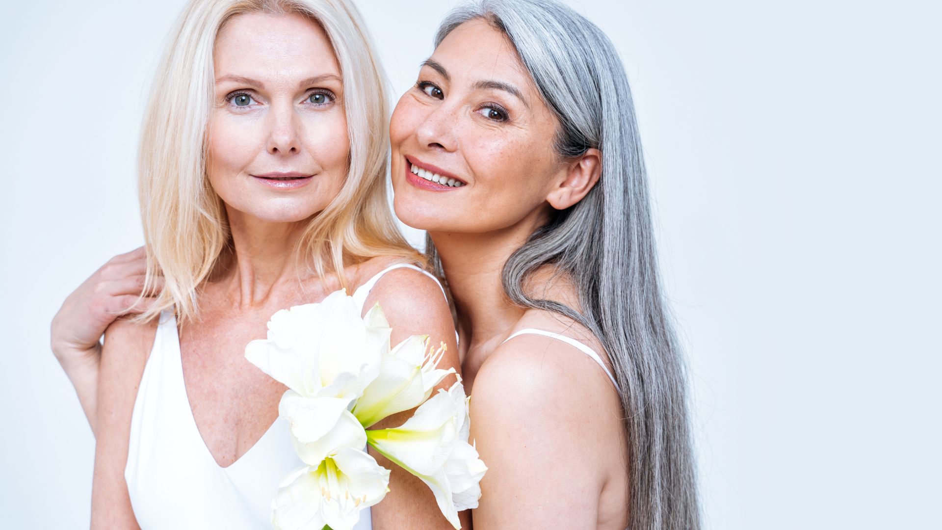 Two women with light complexions, one with blonde hair holding white flowers and the other with long gray hair, smiling at the camera against a light background.