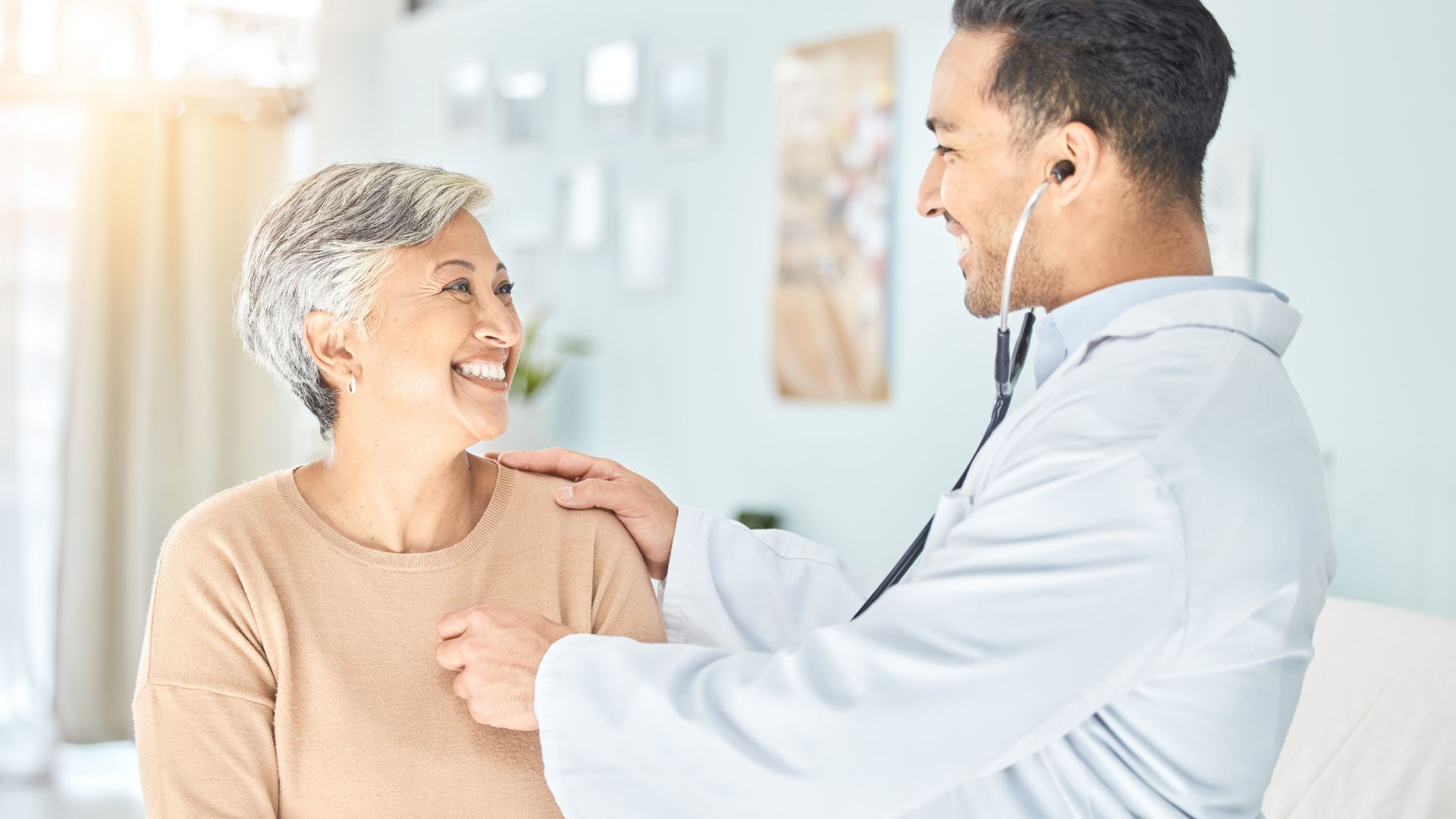 A doctor uses a stethoscope to listen to the chest of an older woman, who is smiling and sitting in a medical office.