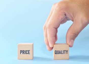 A hand is picking up a wooden block labeled "QUALITY" from a set of two blocks. The other block is labeled "PRICE." The blocks are set against a light blue background.