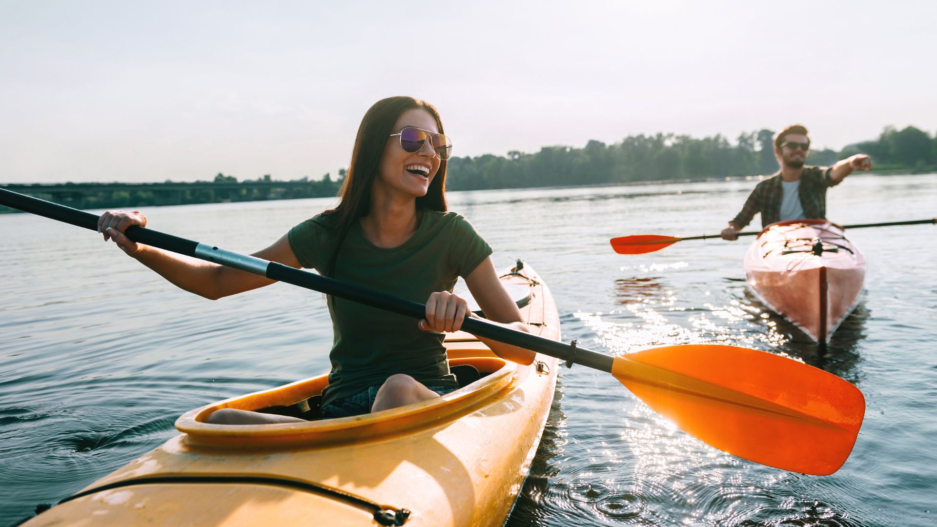 Two individuals kayaking on a calm lake, both are smiling and wearing casual clothing. The person in the foreground is in a yellow kayak, and the person in the background is in an orange kayak.