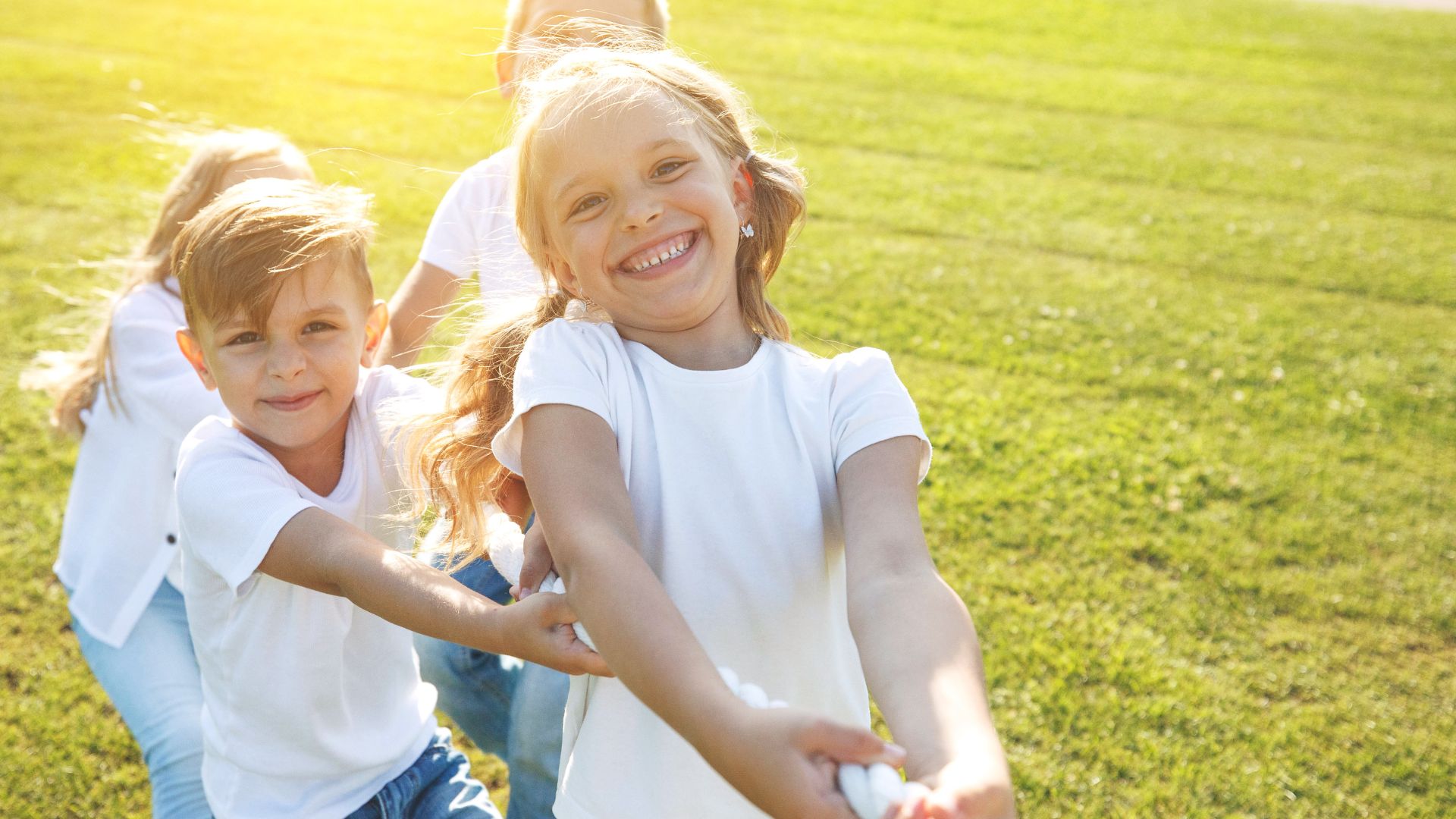 Children smiling and playing tug-of-war on a sunny day in a grassy field.