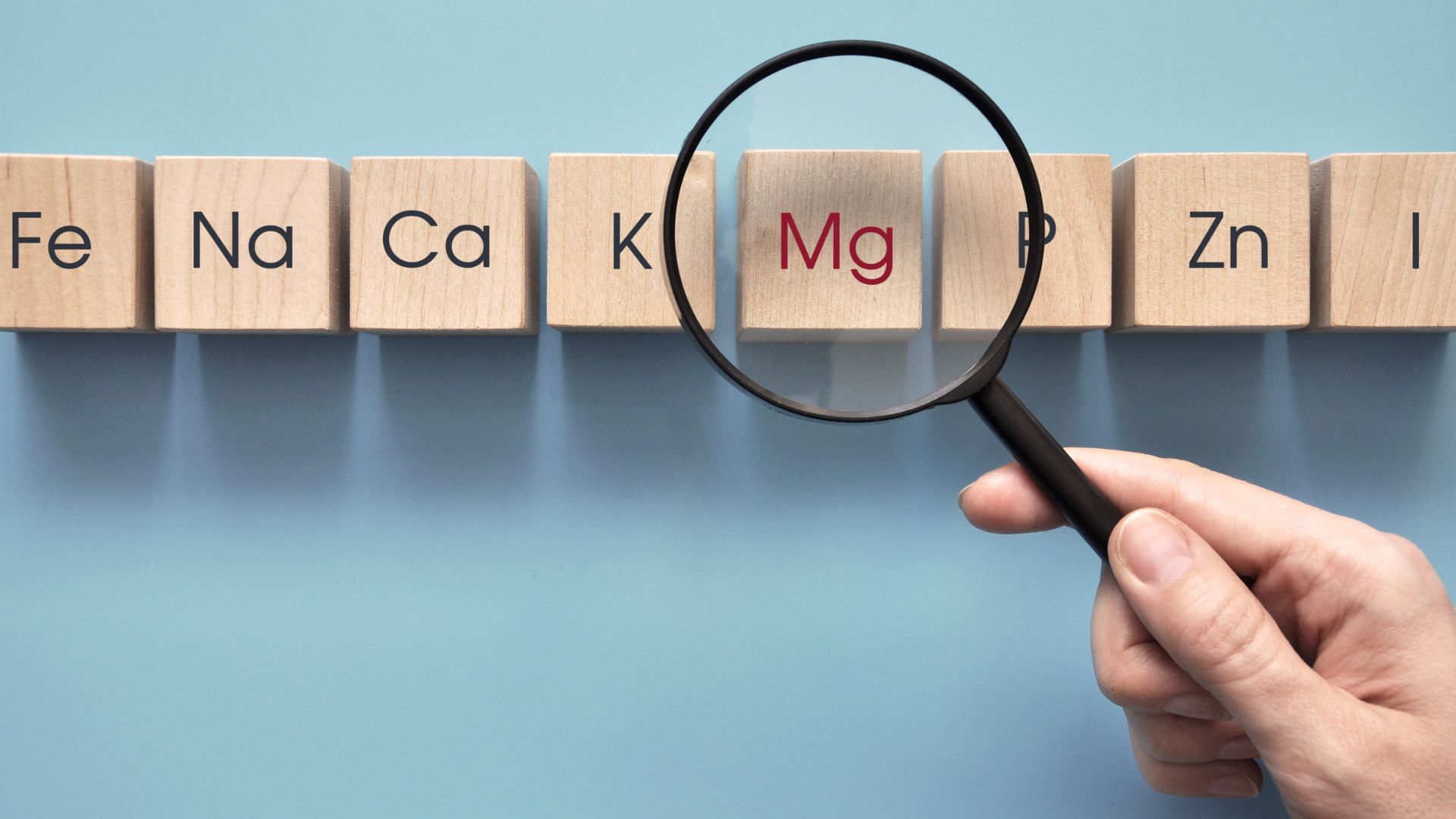A hand holds a magnifying glass over wooden blocks with chemical element symbols, highlighting "Mg" on a blue background.