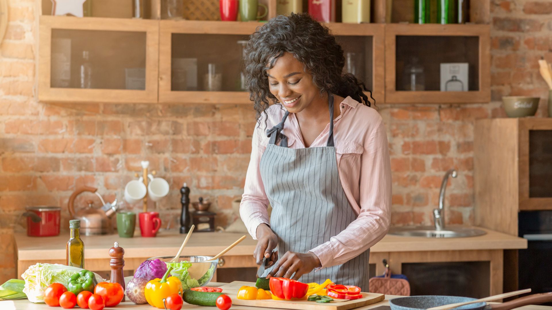 A person in an apron chops vegetables in a kitchen with wooden cabinets and various kitchen items around.
