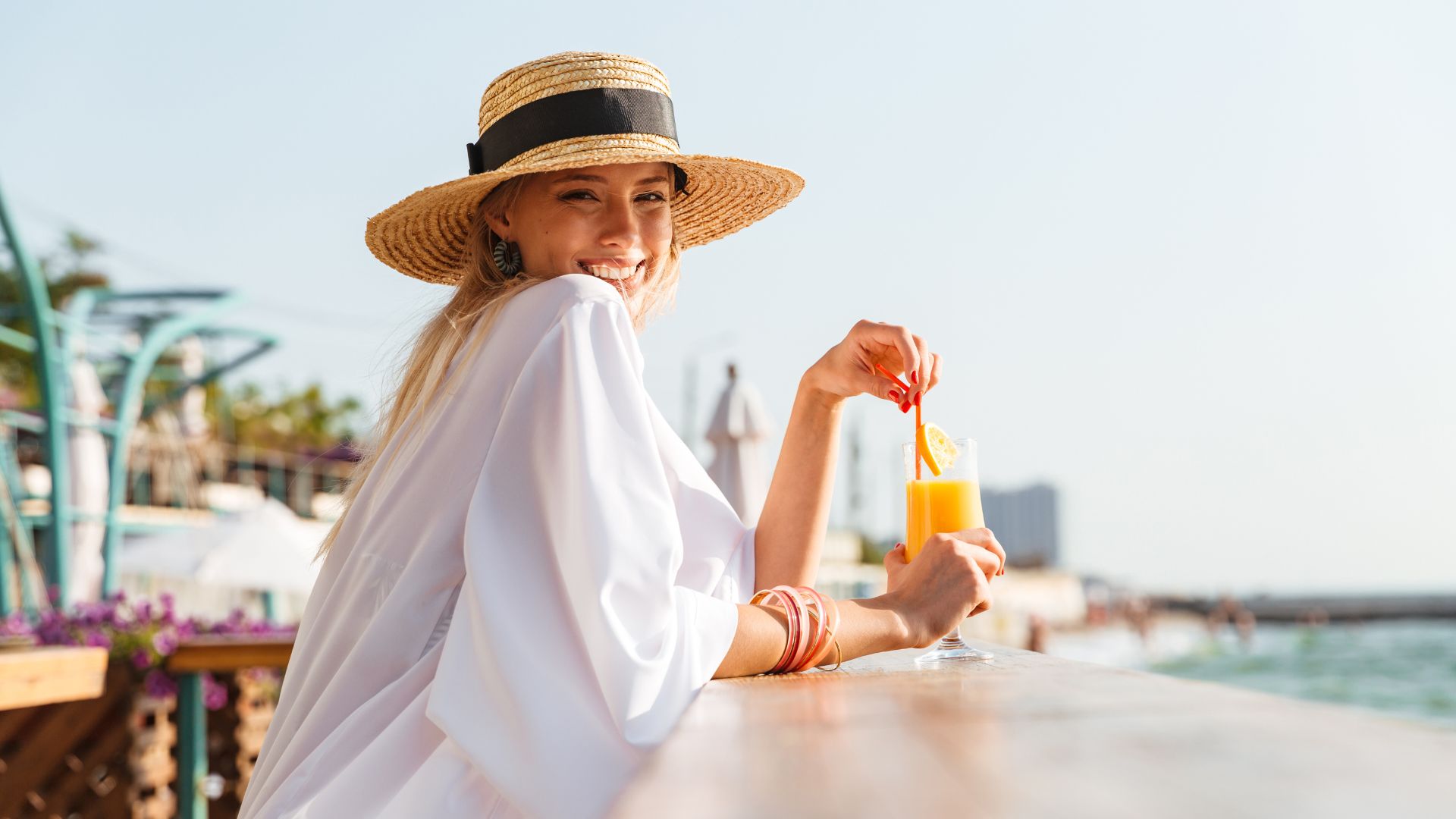 Woman in a white dress and straw hat smiles while sitting by the water, holding a glass of orange juice with a slice of orange on the rim.