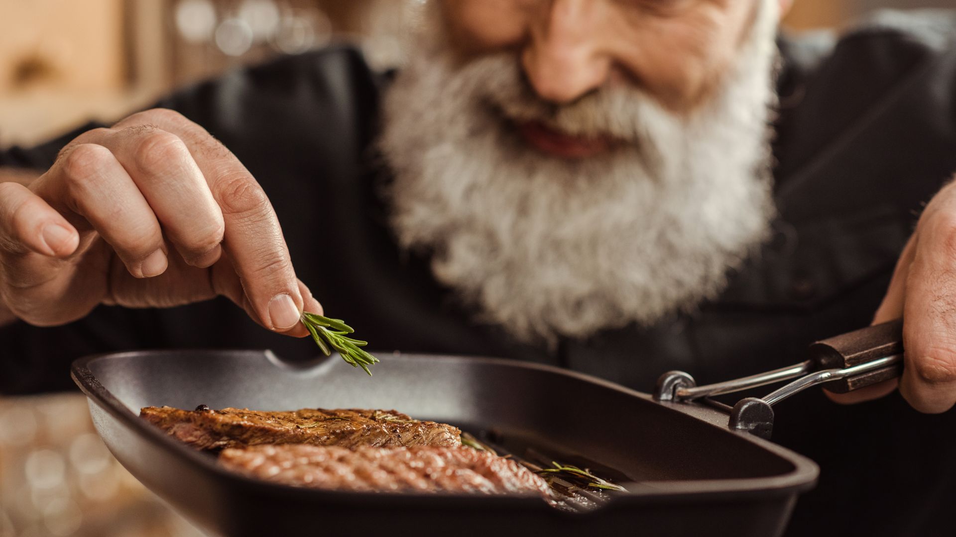 A person with a beard adds a sprig of rosemary to a piece of meat in a frying pan.