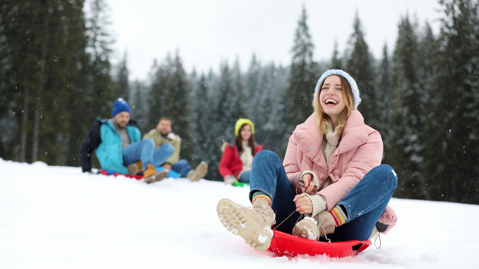 A woman in a pink jacket sleds down a snowy hill, smiling, with three people sledding behind her. Pine trees are in the background.