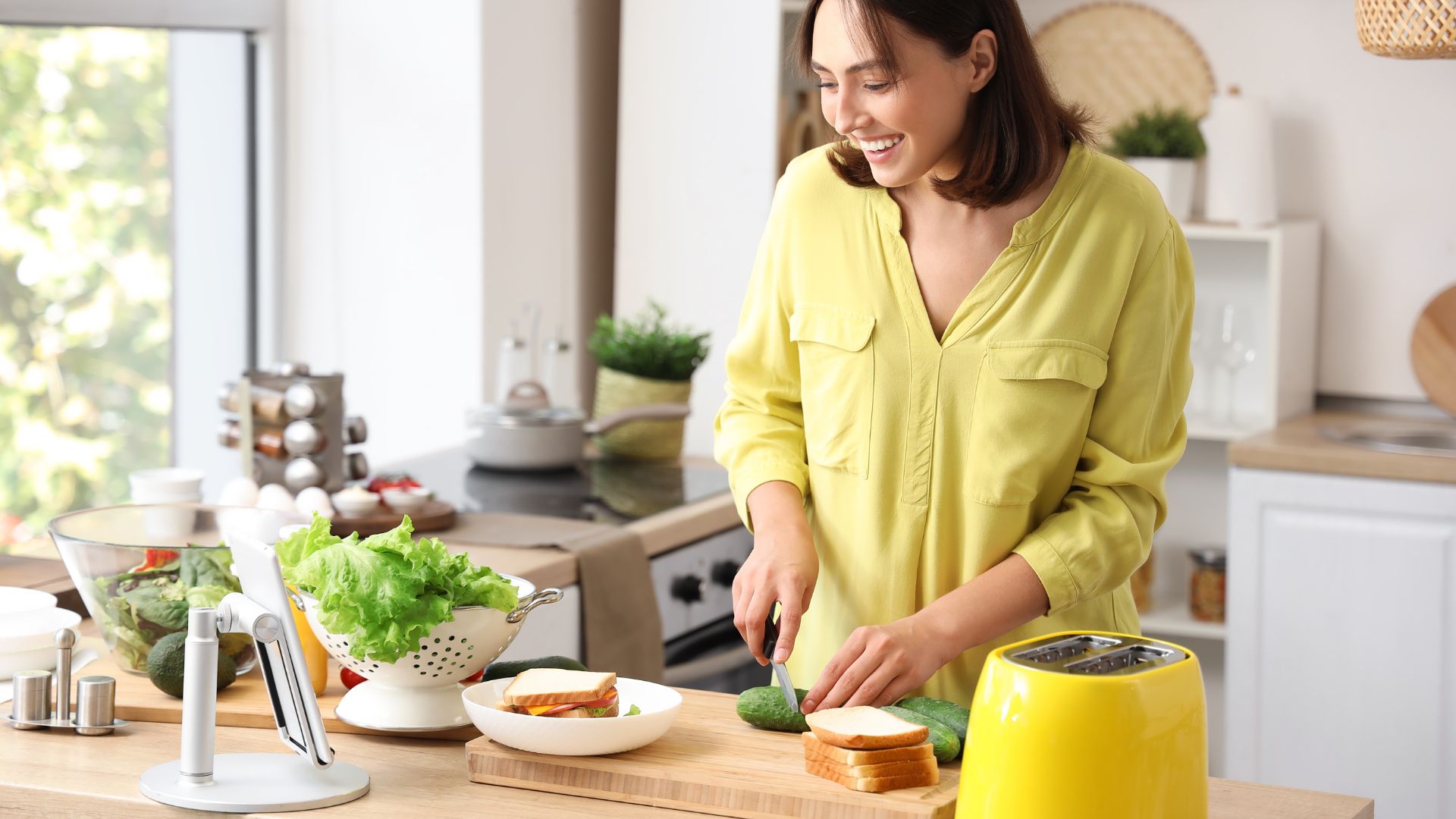 Woman in a yellow shirt slices cucumbers in a bright kitchen, with bread, lettuce, and a yellow toaster nearby.