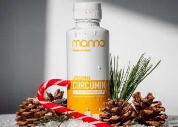 A bottle of Manna Liposomal Curcumin surrounded by pine cones, pine sprigs, snow, and a red and white candy cane, set against a white background.