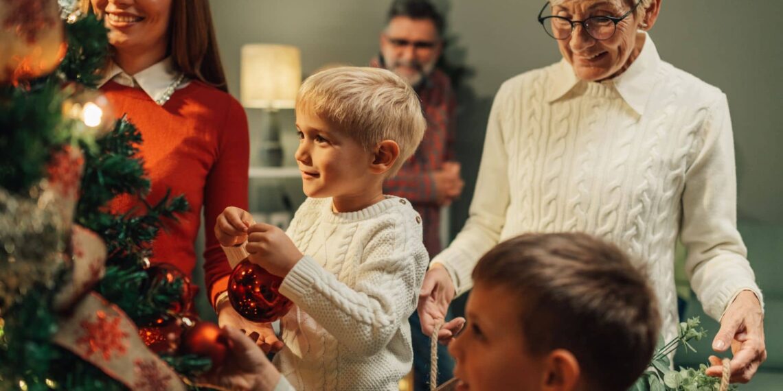 A family decorates a Christmas tree together, with adults and children placing ornaments. One child is holding a red bauble.