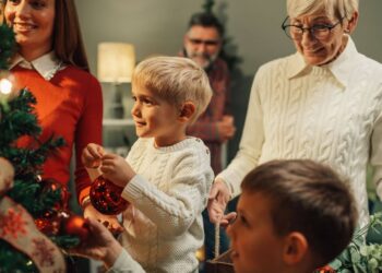 A family decorates a Christmas tree together, with adults and children placing ornaments. One child is holding a red bauble.
