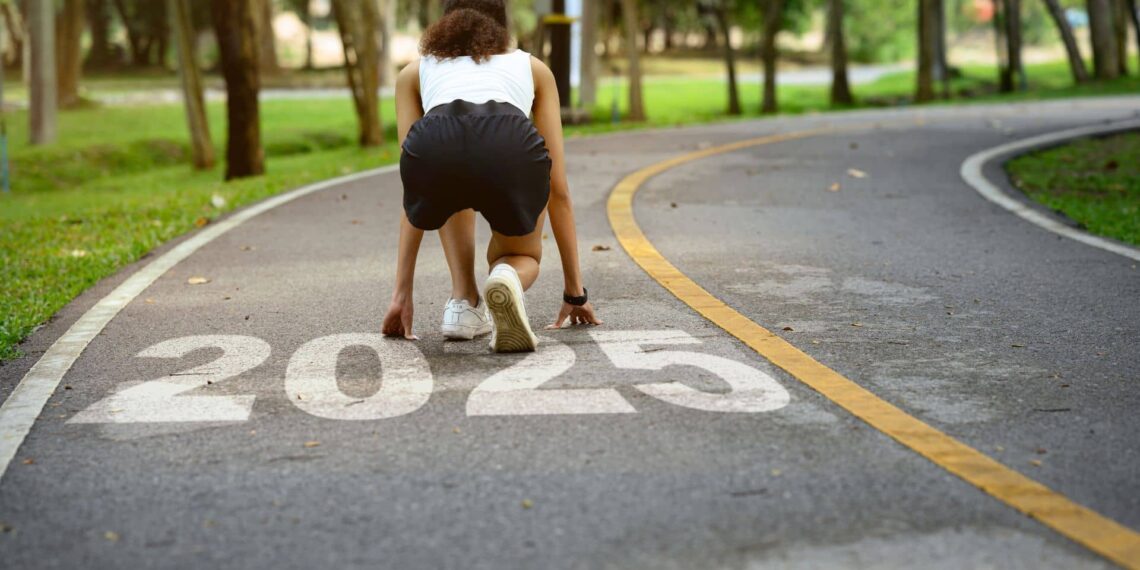 A person in athletic attire is crouched in a starting position on a path with "2025" painted on it, surrounded by trees.