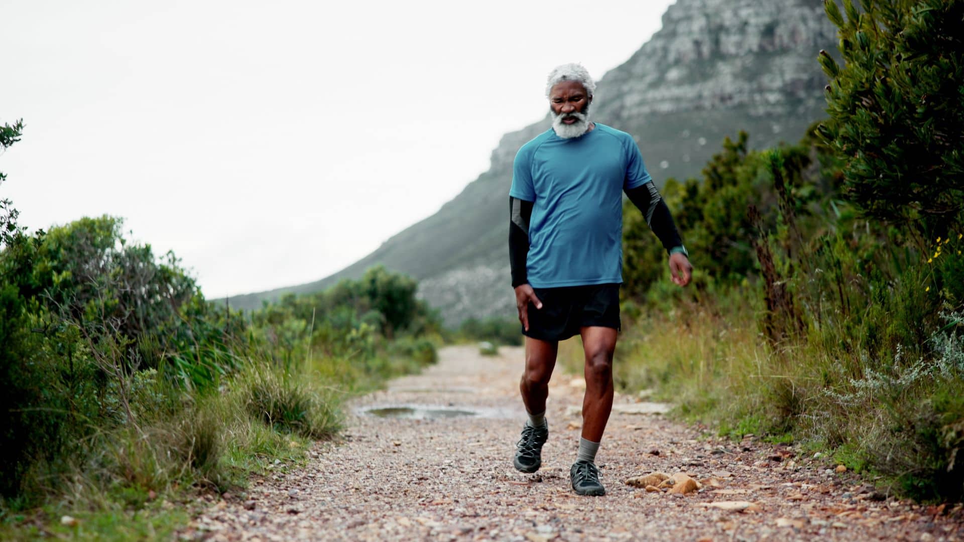 An older man with a gray beard walks along a rocky path in a mountainous, green landscape. He wears a blue shirt and black shorts.