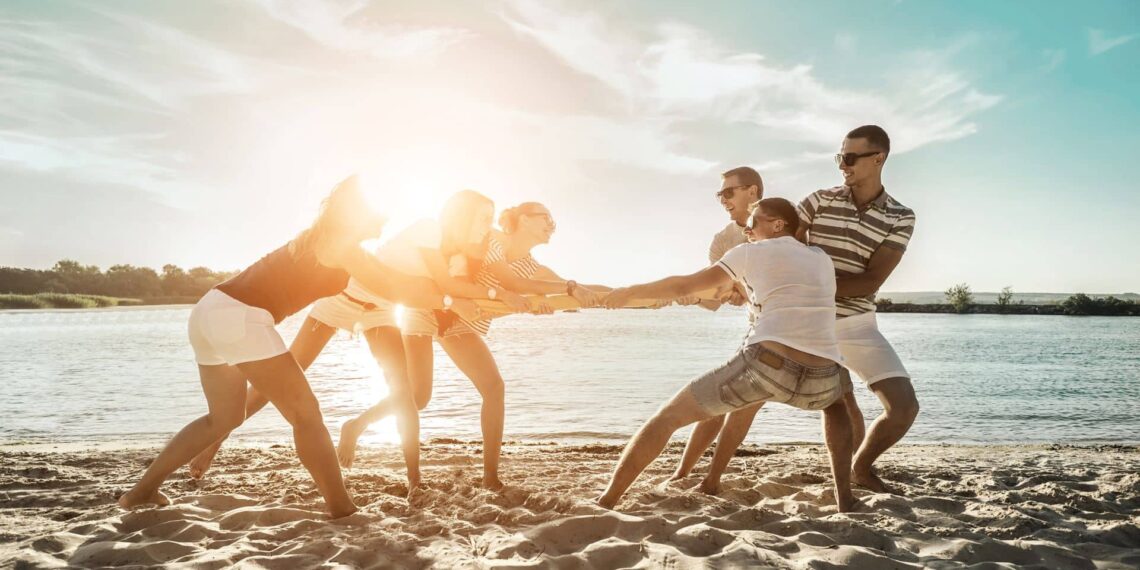 Group of people playing tug-of-war on a sandy beach by the water with the sun shining in the background.