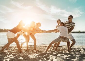 Group of people playing tug-of-war on a sandy beach by the water with the sun shining in the background.