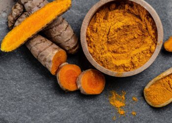 Turmeric roots and powder in a wooden bowl on a dark textured surface.