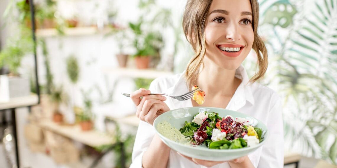 A person smiling while holding a salad bowl and a fork, surrounded by green plants indoors.