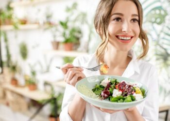 A person smiling while holding a salad bowl and a fork, surrounded by green plants indoors.