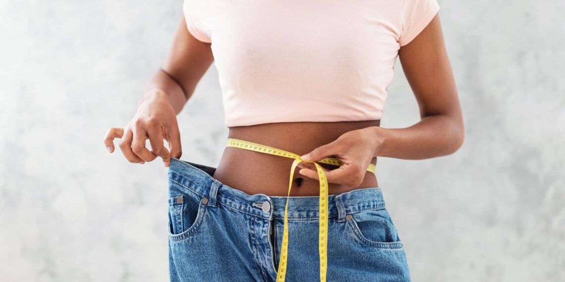 Person in a pink shirt measures their waist with a tape while holding loose-fitting jeans, indicating weight loss.
