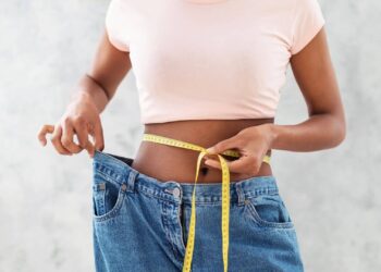 Person in a pink shirt measures their waist with a tape while holding loose-fitting jeans, indicating weight loss.