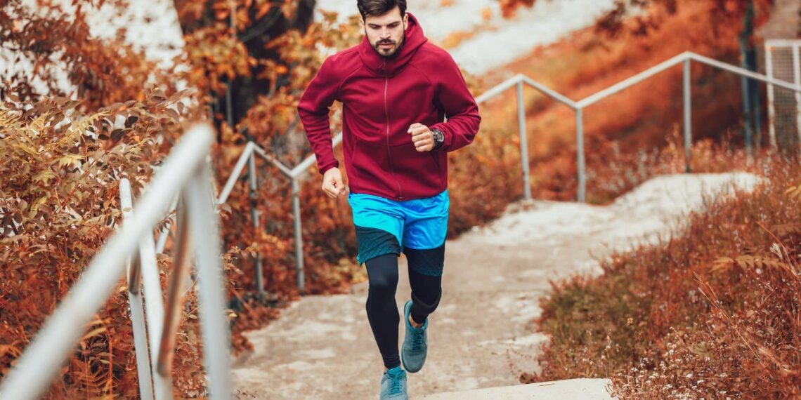 A man in a red hoodie and blue shorts runs up outdoor stairs surrounded by autumn foliage.