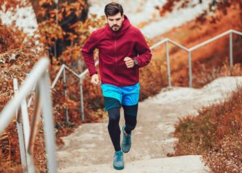 A man in a red hoodie and blue shorts runs up outdoor stairs surrounded by autumn foliage.