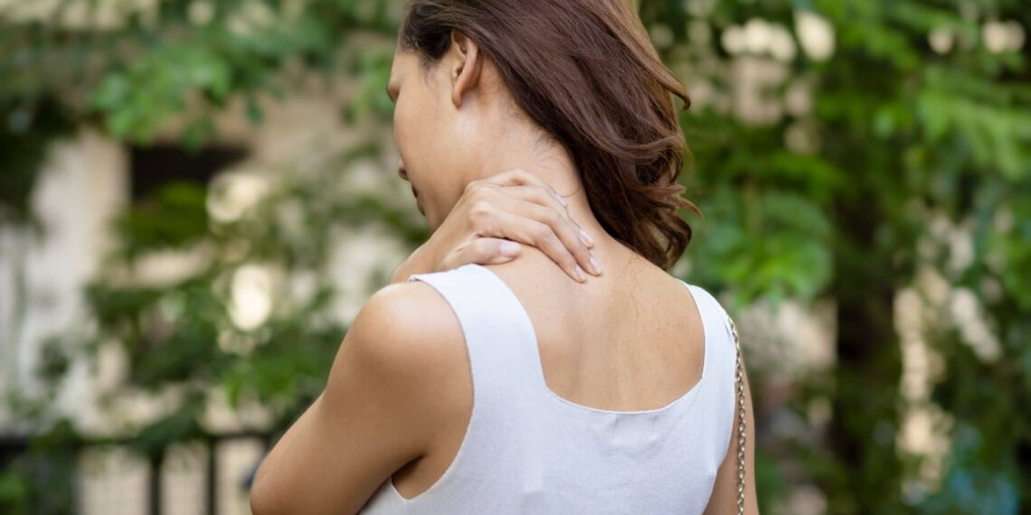 Person in a white top standing outdoors, touching their neck with one hand, surrounded by green foliage.