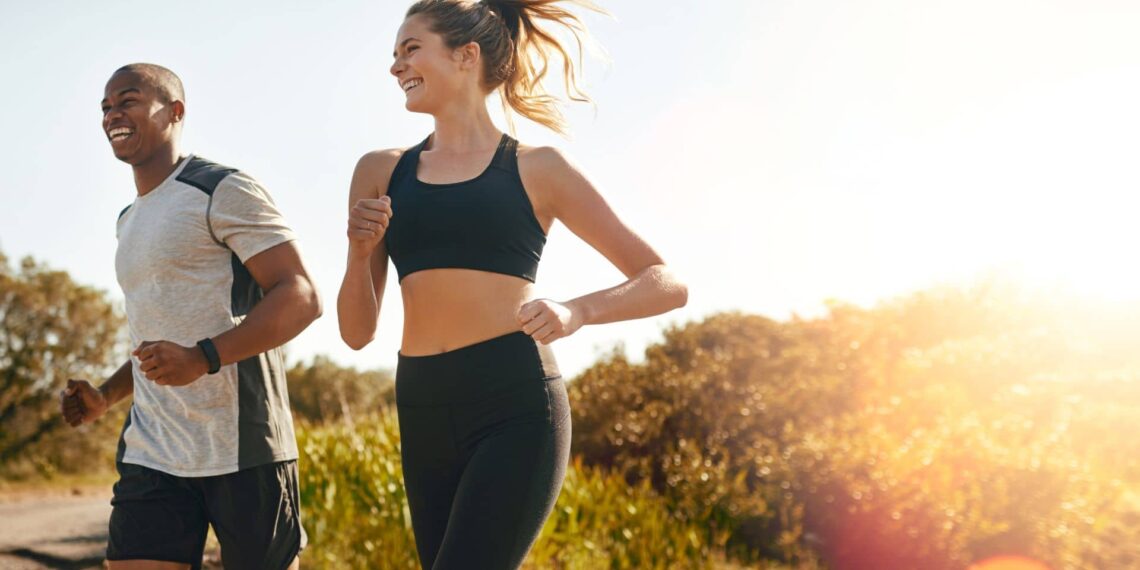 A man and a woman jogging outdoors on a sunny day, with trees and vegetation in the background.